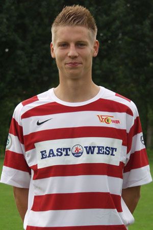 Ein junger Fußballspieler in einem gestreiften Trikot mit dem Schriftzug "EAST WEST", steht lächelnd vor einem grünen Hintergrund.