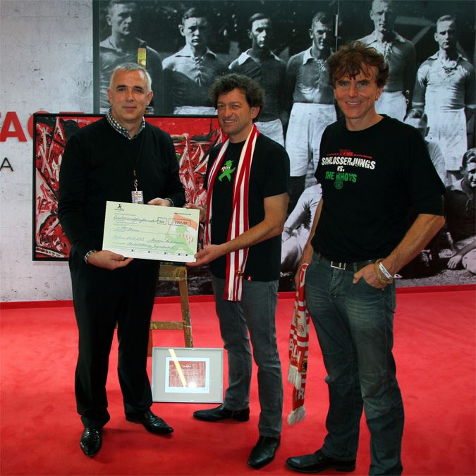 Drei Männer stehen vor einer schwarz-weißen Fotowand, halten einen Scheck und posieren freundlich für die Kamera.