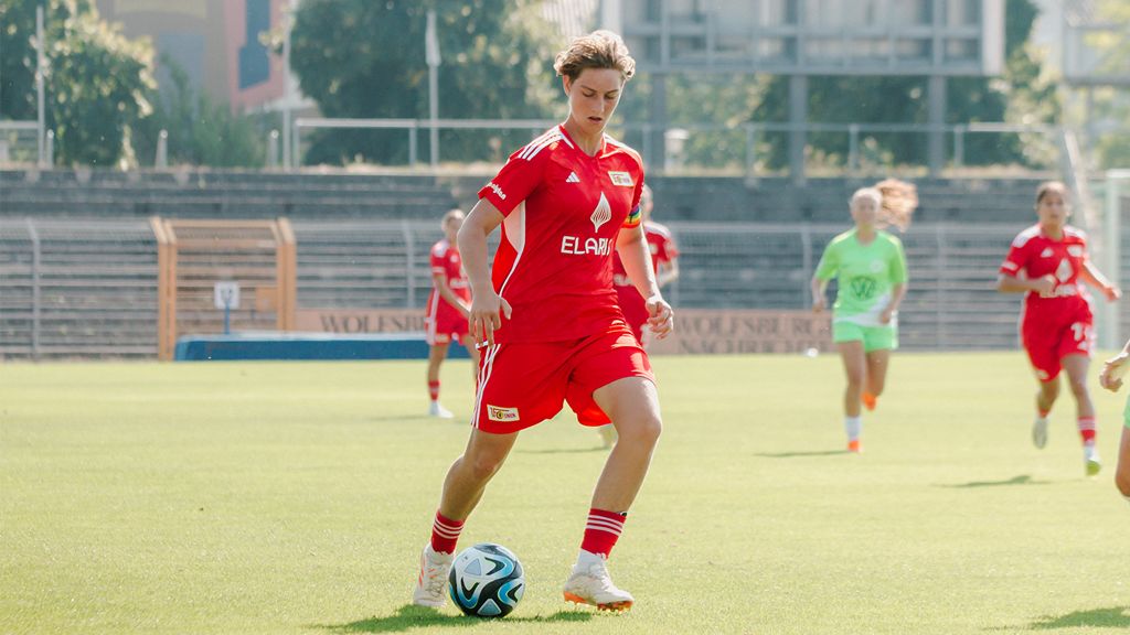 A soccer player in a red jersey dribbles the ball on a training field, while teammates stand in the background.
