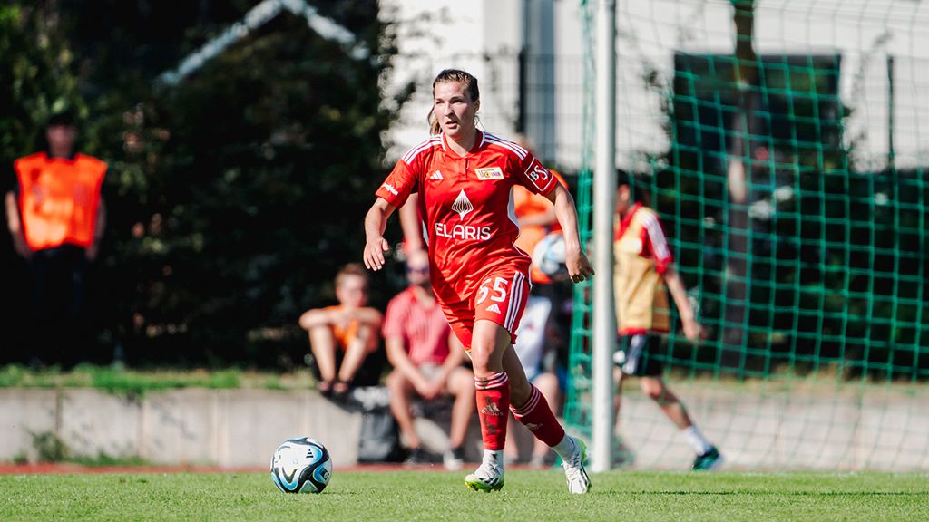 A player in a red jersey with the number 55 is dribbling the ball on the soccer field. Spectators in the background.