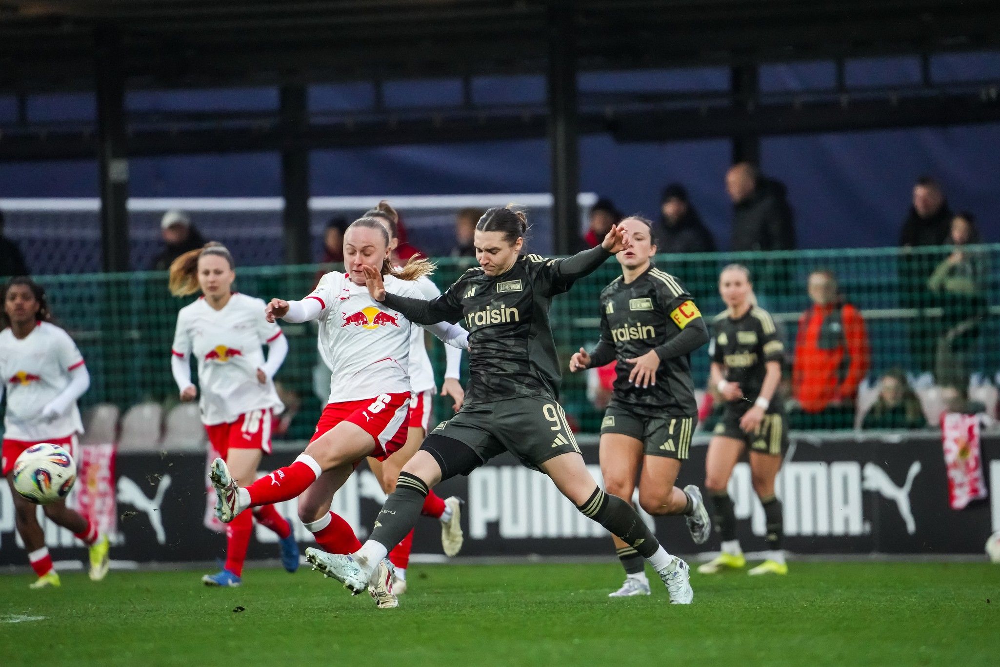 Ein Fußballspiel zwischen zwei Mannschaften, wobei eine Spielerin in Schwarz den Ball von einer Spielerin in Weiß erobert.