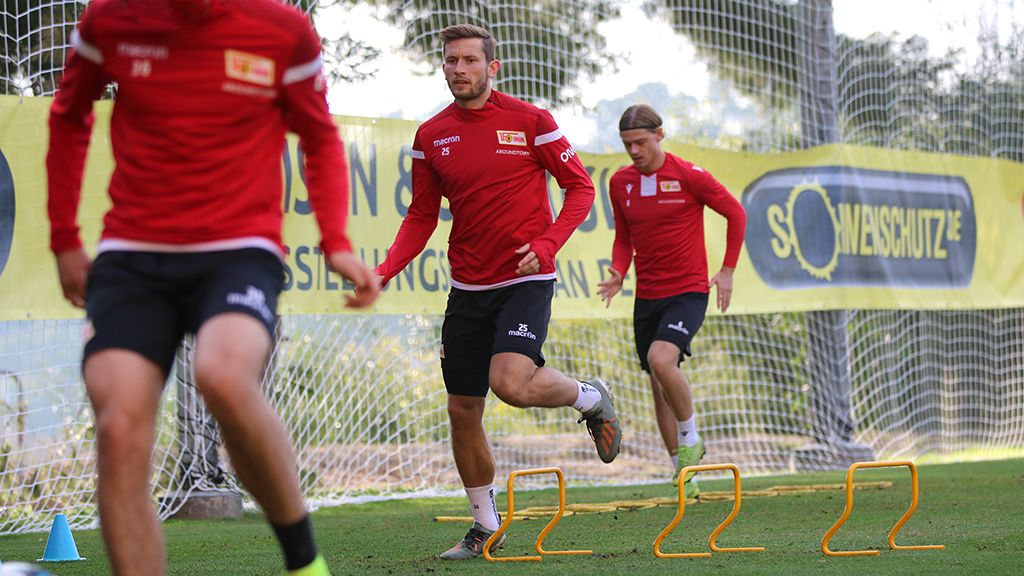 Drei Fußballspieler in roten Trikots trainieren auf einem Rasenplatz, während sie über Hindernisse sprinten.