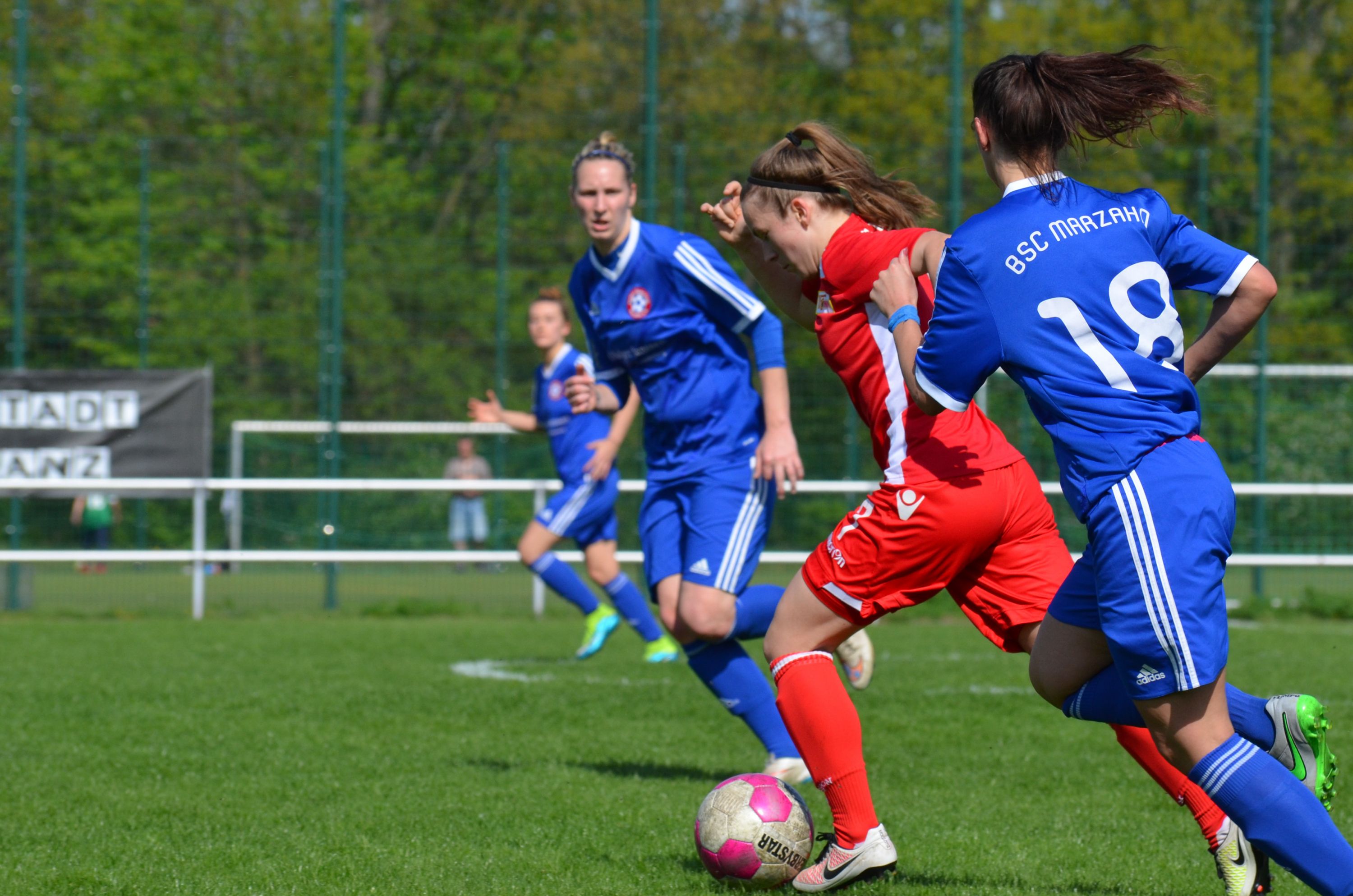 Zwei Fußballspielerinnen in roten und blauen Trikots kämpfen um den Ball auf einem grünen Fußballfeld.
