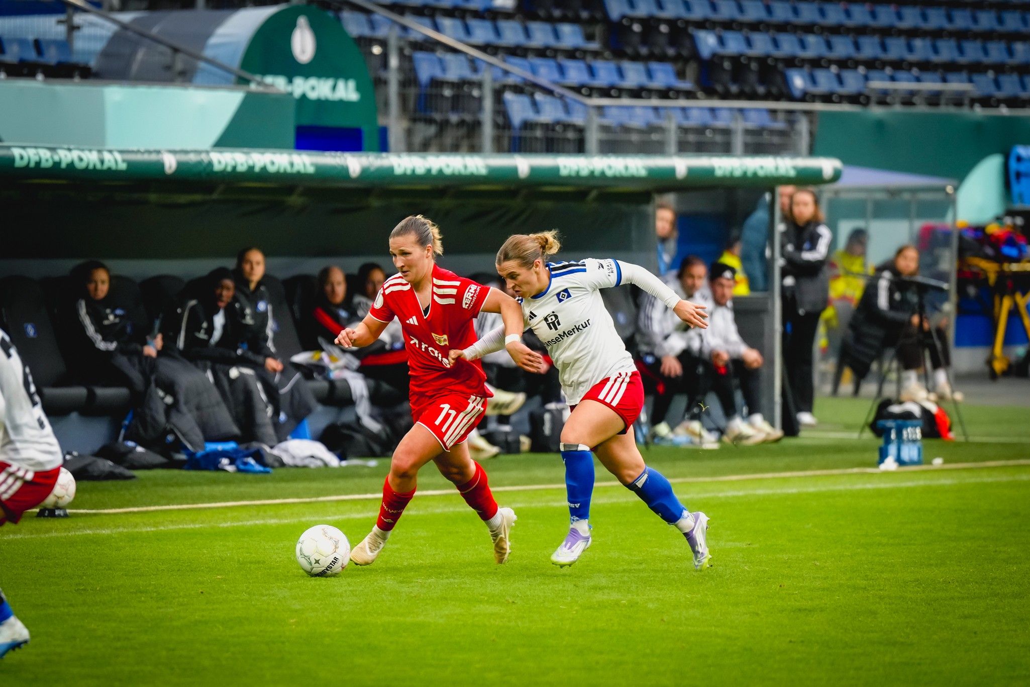 Zwei Fußballspielerinnen in roten und weißen Trikots jagen den Ball auf einem Rasenfeld mit Zuschauerrängen im Hintergrund.