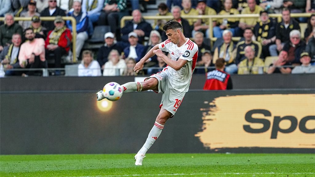 Ein Fußballspieler führt einen Schuss aus, während Zuschauer im Hintergrund zu sehen sind.