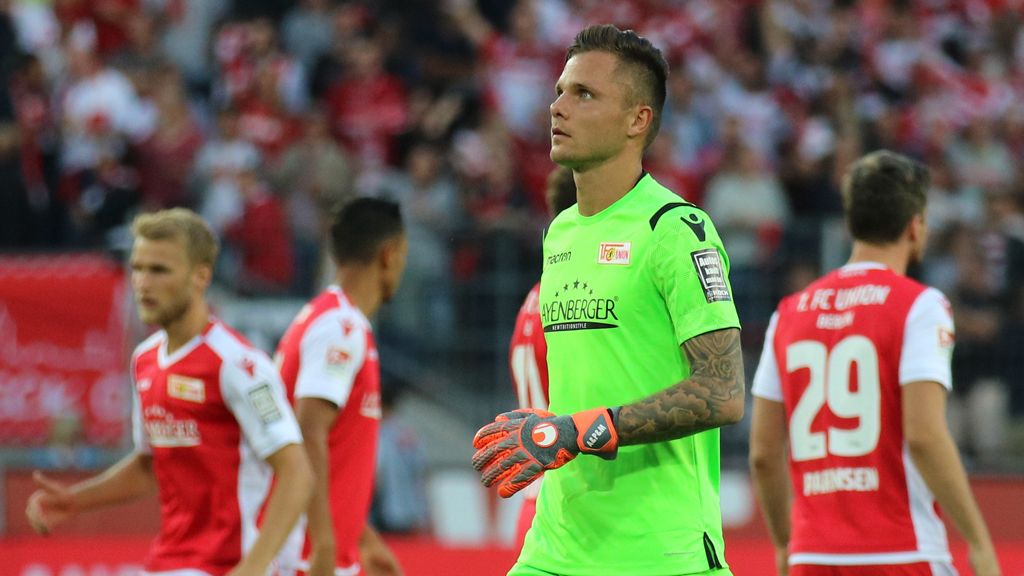 Ein Fußballtorwart in einem grünen Trikot mit Handschuhen, während Mitspieler im Hintergrund mit enttäuschten Mienen stehen.