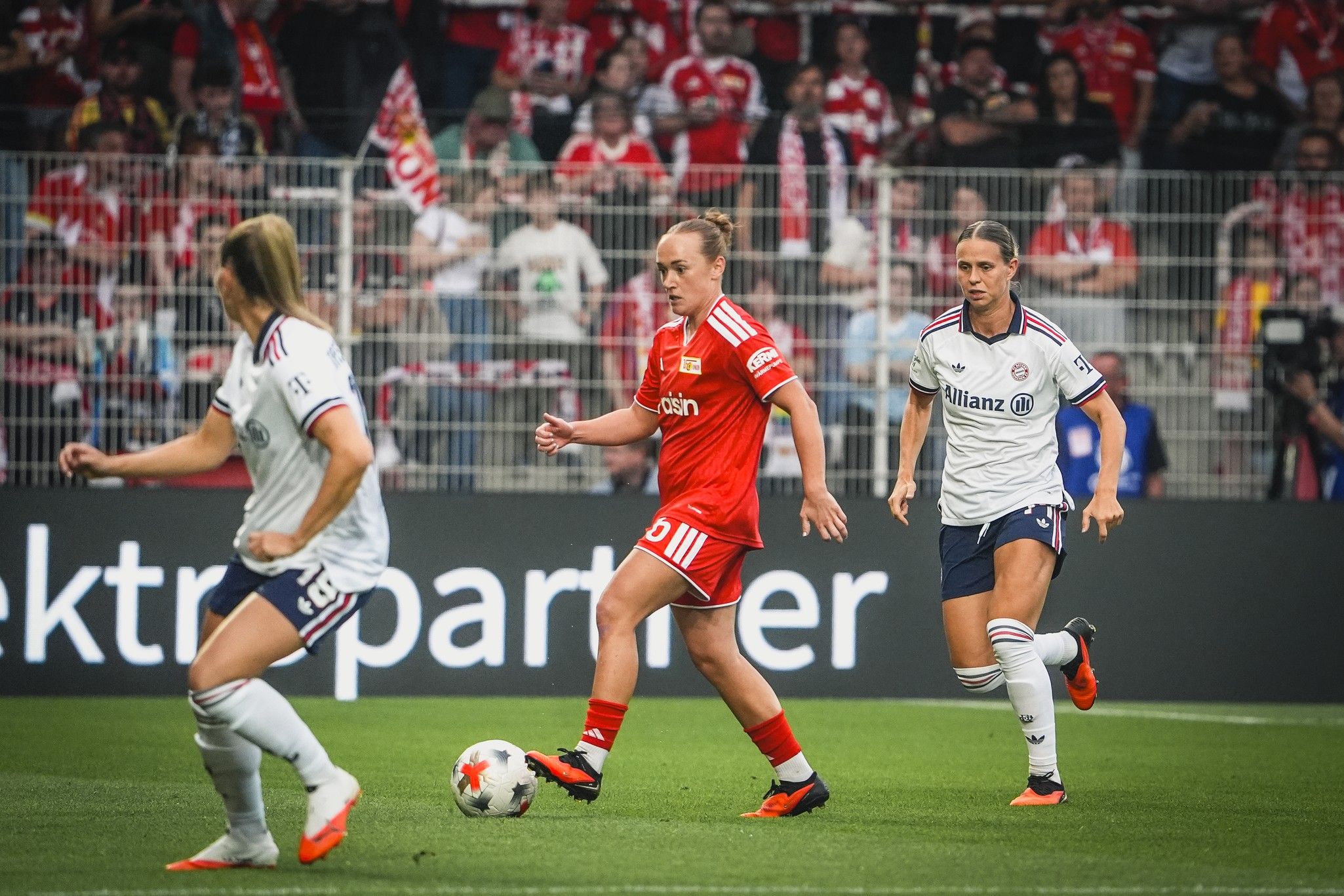Eine Fußballspiel-Szene mit zwei Spielerinnen in weißen Trikots und einer in rotem Trikot, die den Ball spielt, im Stadion.