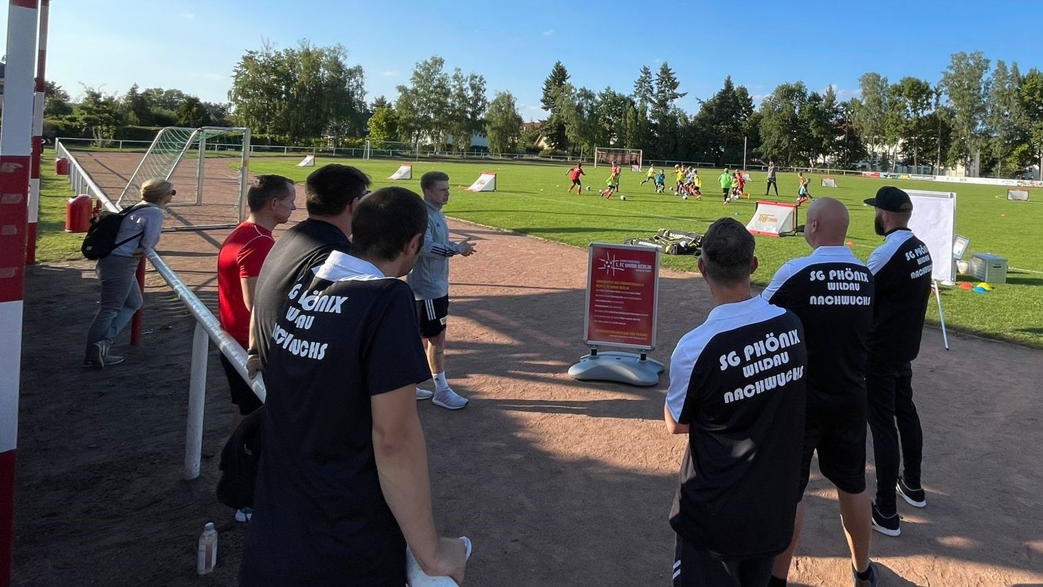 Gruppe von Erwachsenen in Sportkleidung beobachtet eine Trainingssession auf einem Fußballplatz.