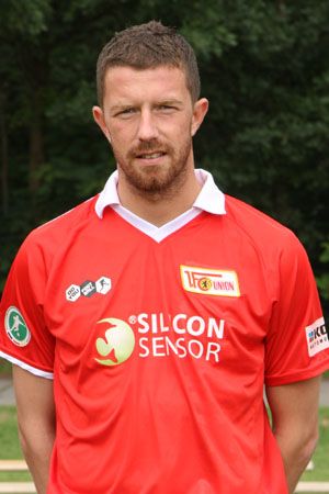 Mann in rotem Fußballtrikot mit Vereinslogo, steht vor einem grünen Hintergrund.