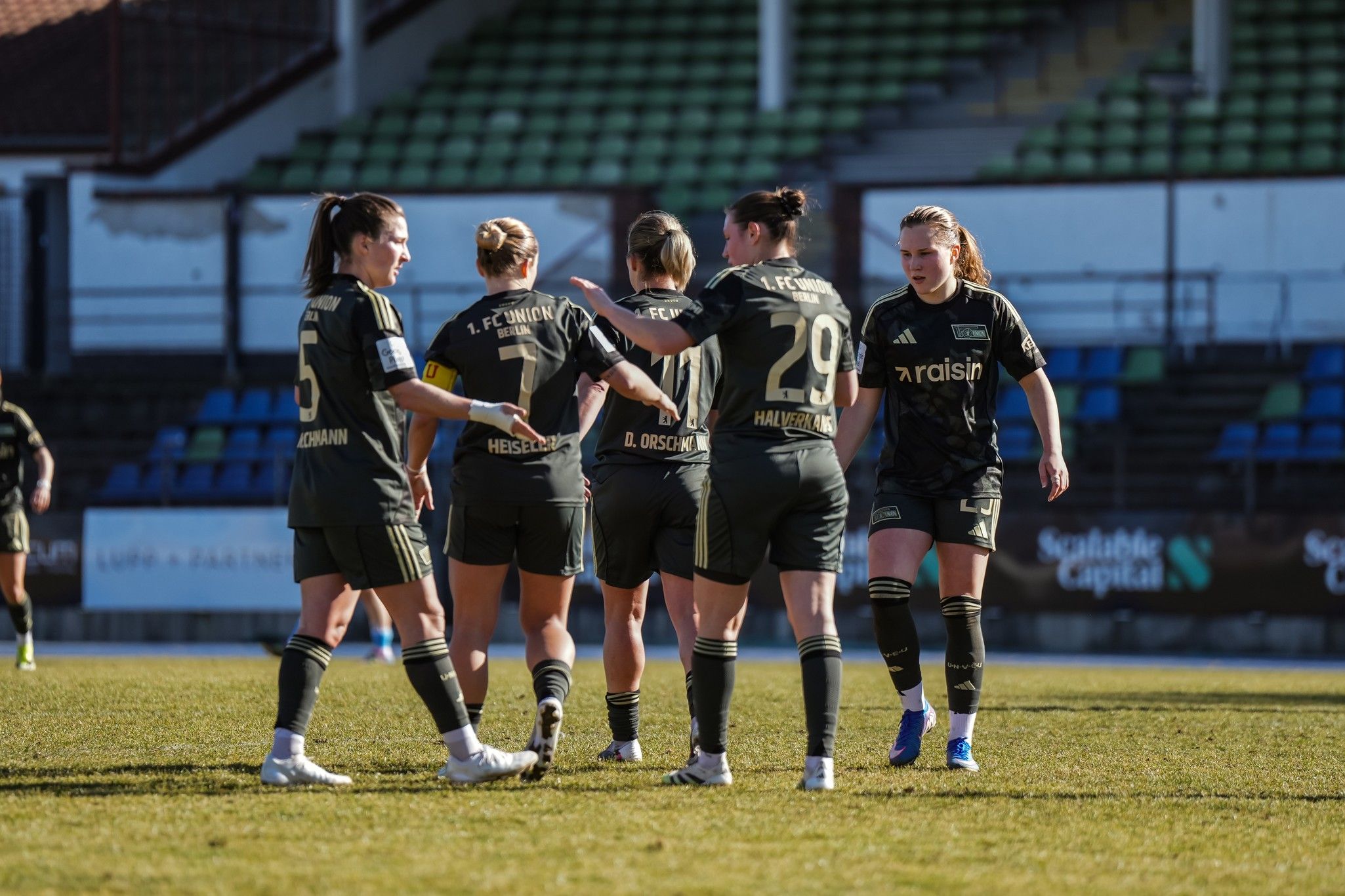 Fünf Frauen in schwarzen Fußballtrikots stehen auf einem Spielfeld und feiern gemeinsam nach einem Spielzug.