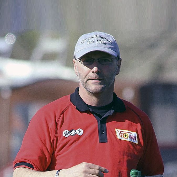 Mann mit Brille und Baseballkappe in rotem Polo-Shirt, steht im Freien und hält eine Flasche.