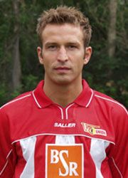 Ein junger Mann in einem roten Fußballtrikot mit weißen Streifen und einem Sponsorenlogo steht vor einer unscharfen grünen Kulisse.