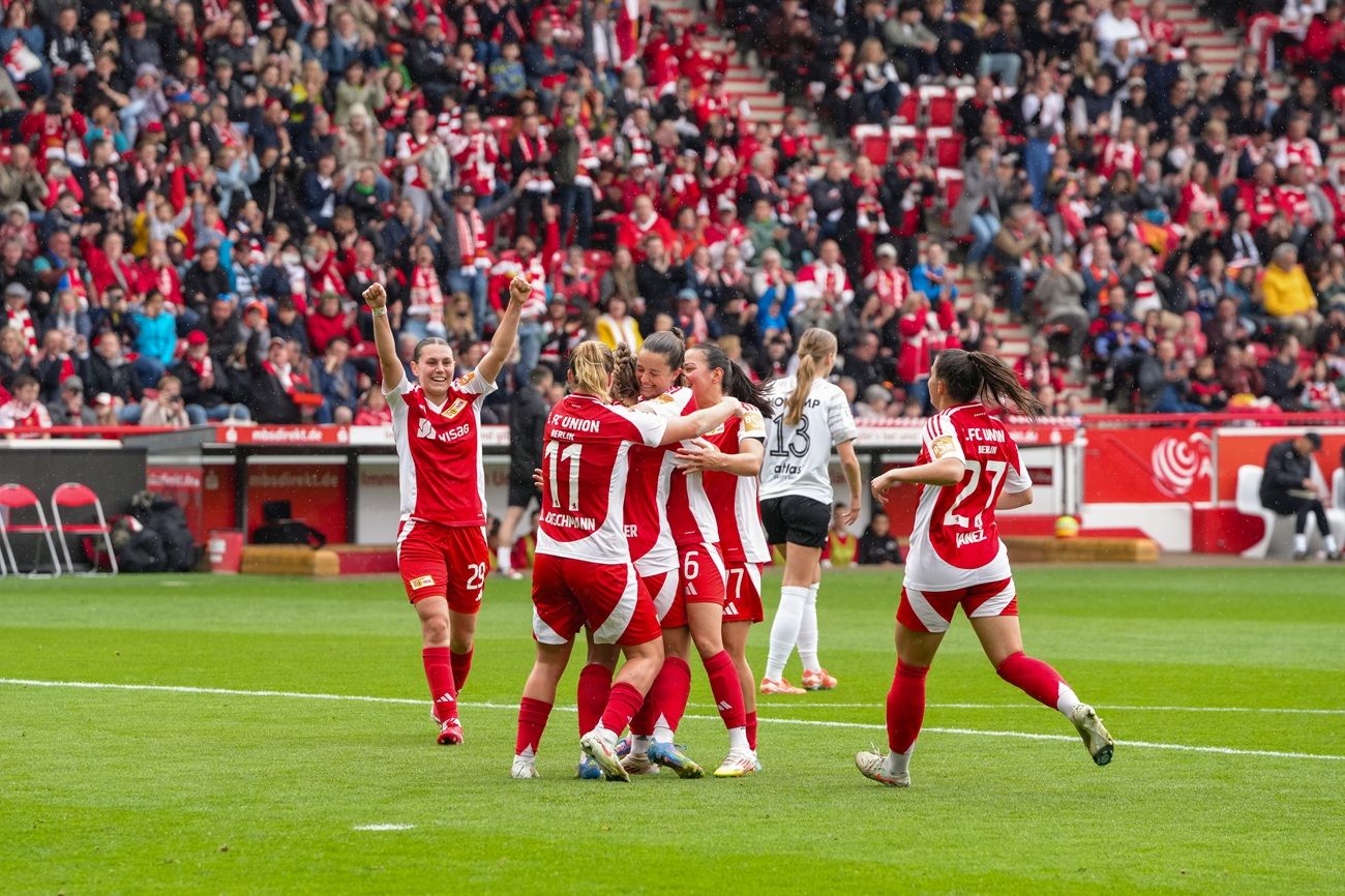 Spielerinnen einer Fußballmannschaft feiern ein Tor vor jubelndem Publikum in einem Stadion.