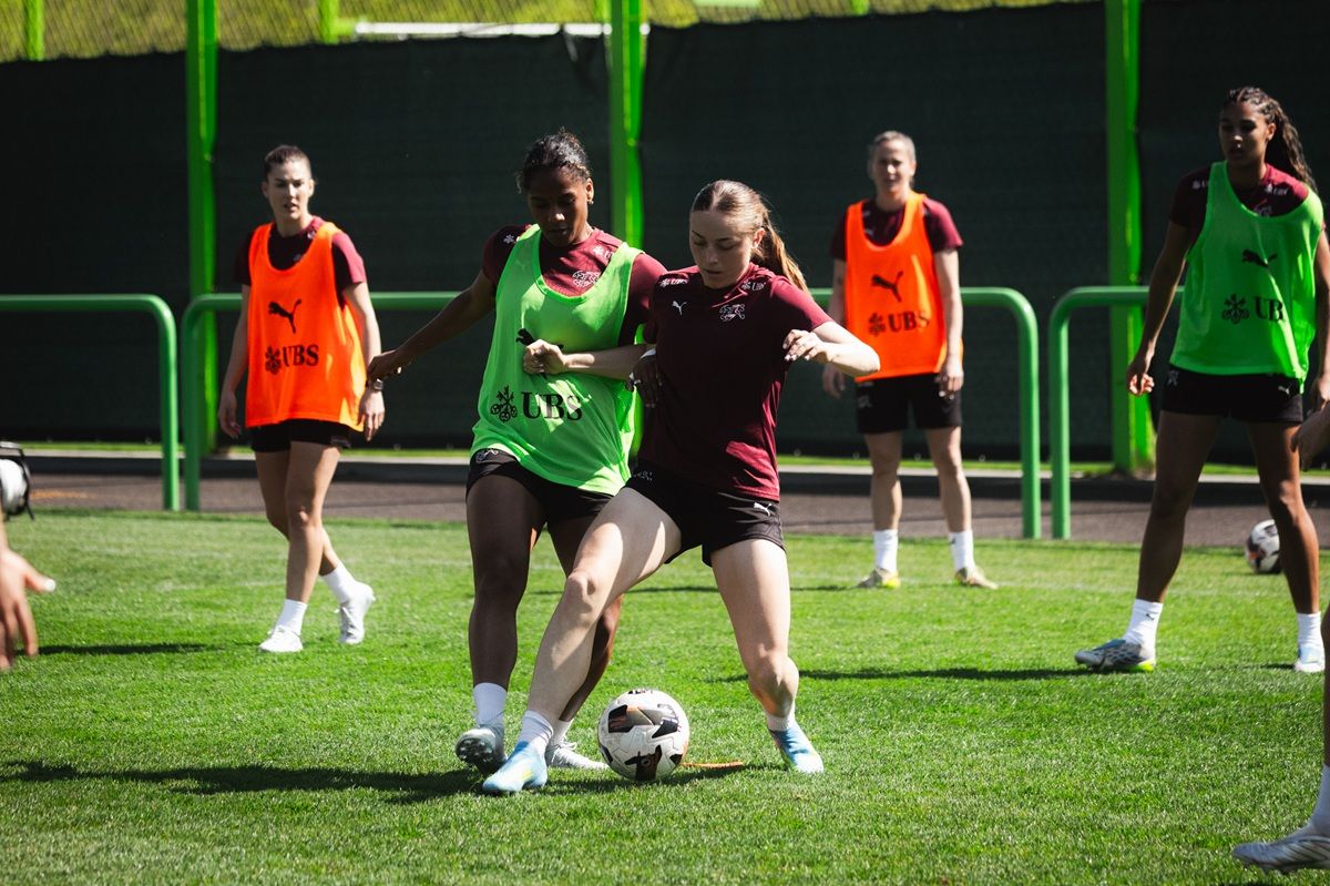 Spielerinnen trainieren auf einem Fußballplatz, während eine Spielerin den Ball dribbelt und eine andere sie verfolgt.