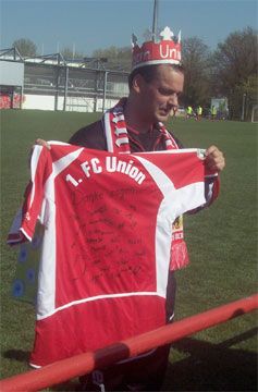 Ein Mann in einem roten und weißen Fußballtrikot hält ein signiertes Trikot des 1. FC Union und trägt eine Krone.