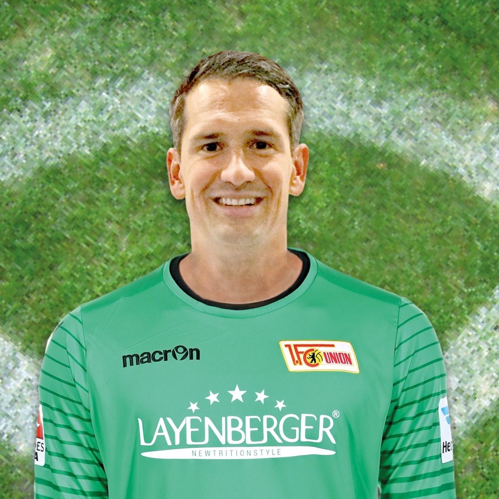 Ein Torwart in einem grünen Trikot mit Sponsorenschriftzug, lächelt vor einem unscharfen, grünen Hintergrund.