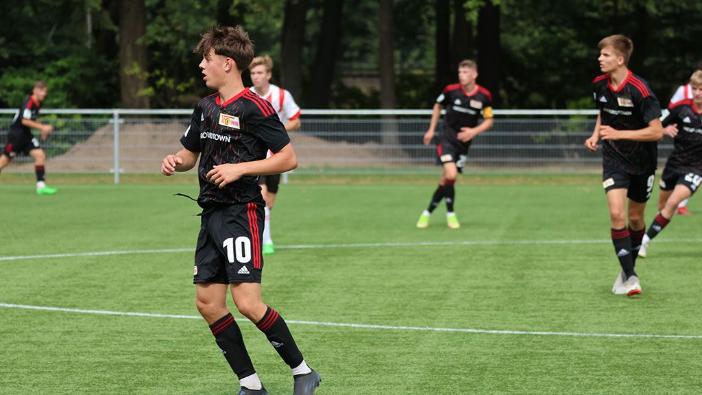 Junge Fußballspieler in Trikots mit Vereinslogo auf einem grünen Spielfeld, einige andere Spieler im Hintergrund.