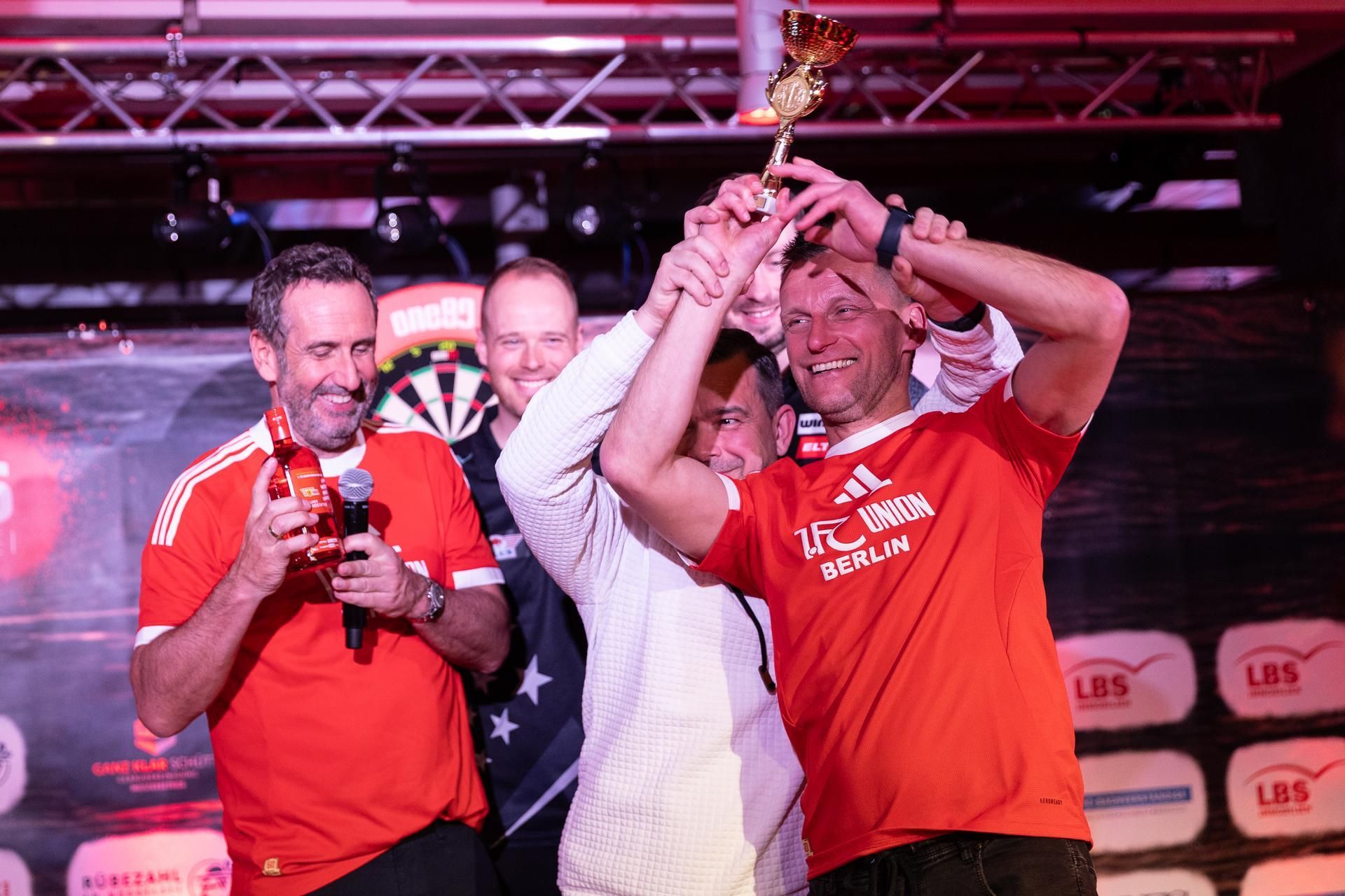 Ein feierlicher Moment: Ein Mann in rotem Shirt von Union Berlin hält einen Pokal, umgeben von drei weiteren Personen.