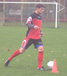 Ein Fußballspieler in rot-schwarzer Trainingskleidung dribbelt einen Ball um ein Hütchen auf einem grünen Platz.