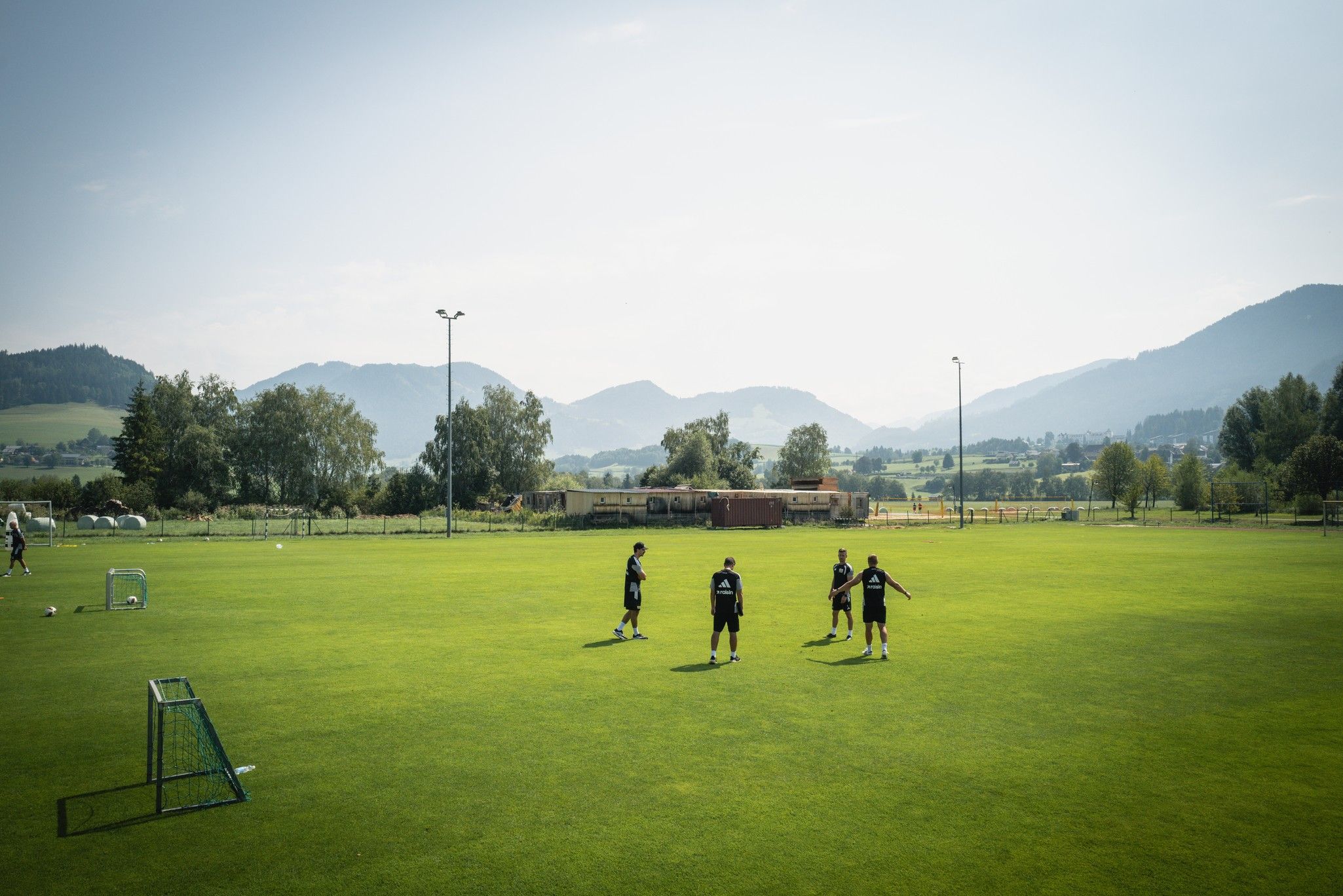 Vier Personen trainieren auf einem grünen Fußballfeld in einer ländlichen Umgebung mit Bergen im Hintergrund.