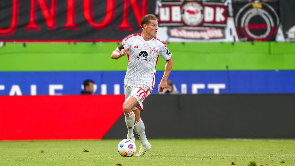 Ein Fußballspieler in einem weißen Trikot mit roten Akzenten dribbelt den Ball auf einem Spielfeld, während Fans im Hintergrund Banner halten.