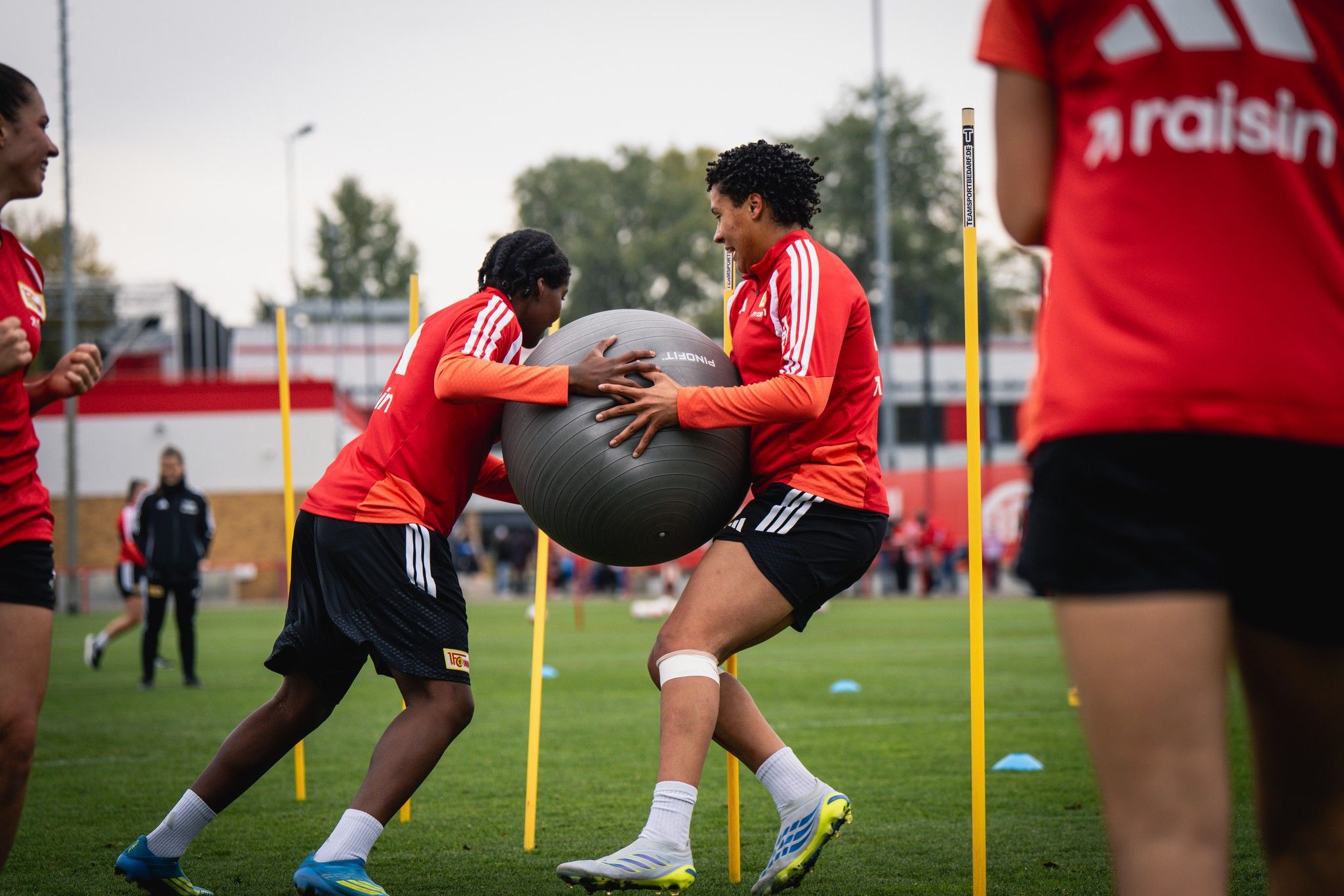 Zwei Sportlerinnen in roten Trainingsanzügen üben mit einem Gymnastikball auf einem Fußballfeld.