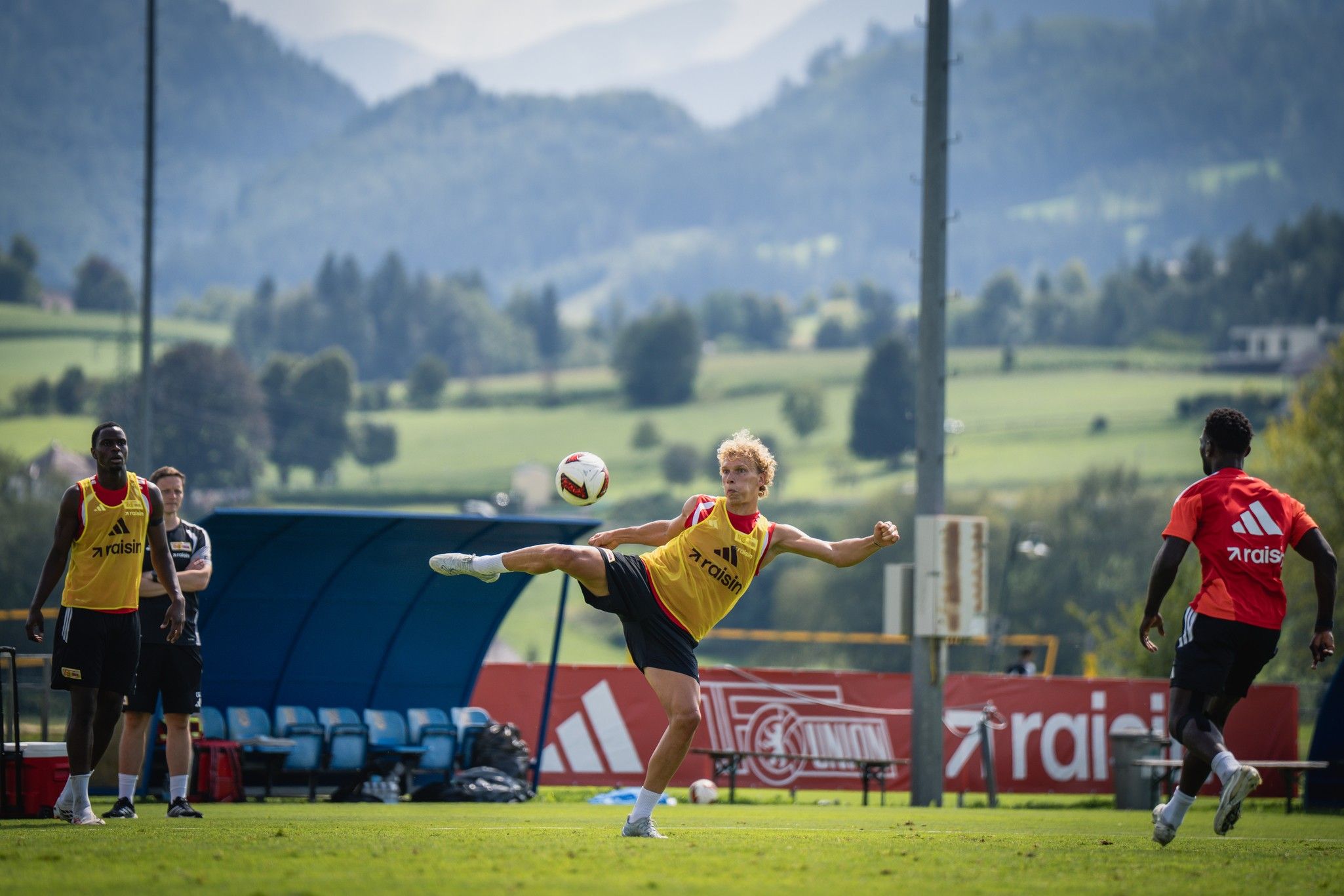 Ein junger Fußballspieler führt einen Schuss mit dem rechten Fuß auf einem Trainingsplatz aus, umgeben von Natur und weiteren Spielern.