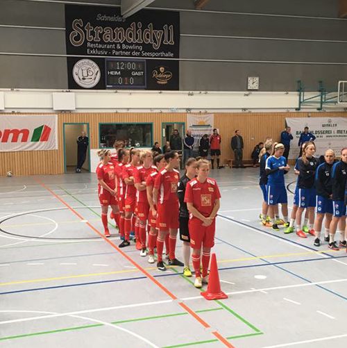 Zwei Fußballmannschaften stehen aufgestellt in einer Sporthalle, eine in roten und die andere in blauen Trikots.