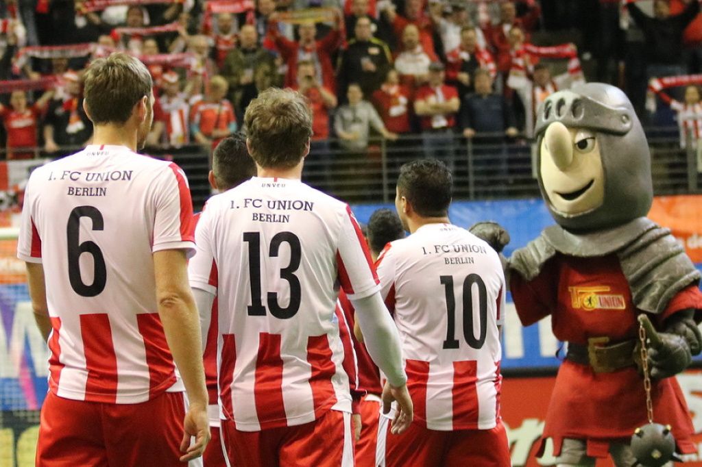 Spieler des 1. FC Union Berlin in Trikots vor einer Menschenmenge, neben einem Maskottchen in Ritterkostüm.