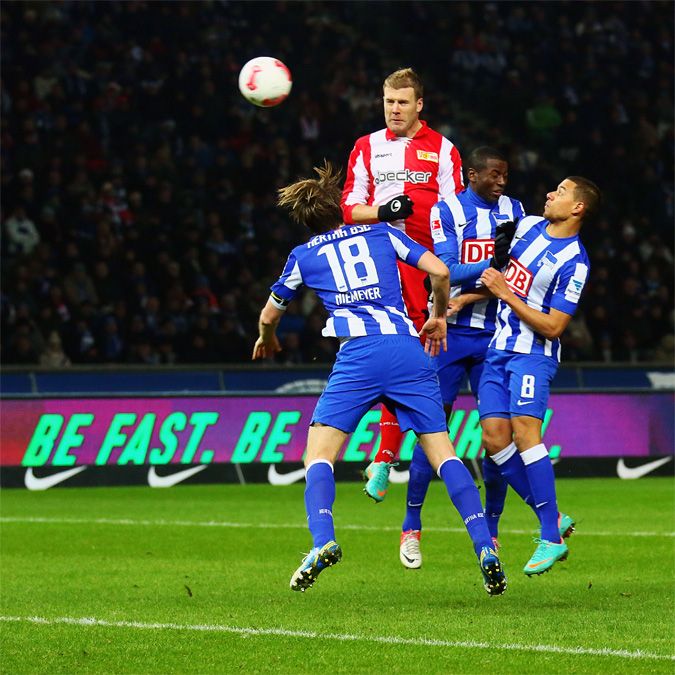 Spielszene im Fußball, Spieler in blauen und roten Trikots springen zum Kopfball in einem Stadion.