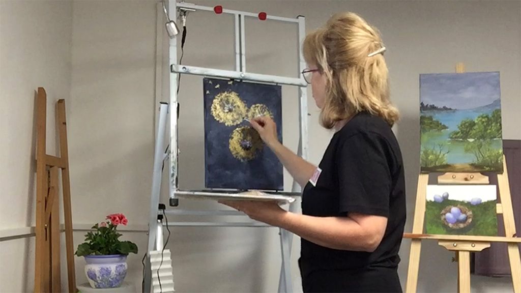 Eine Frau malt an einer staffelei mit zwei runden Bildern. Im Hintergrund ist ein weiteres Gemälde und eine Pflanze auf einem Tisch zu sehen.