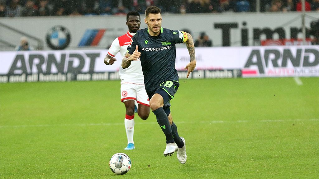 Game scene with a soccer player in blue team gear, dribbling the ball while an opposing player chases him.