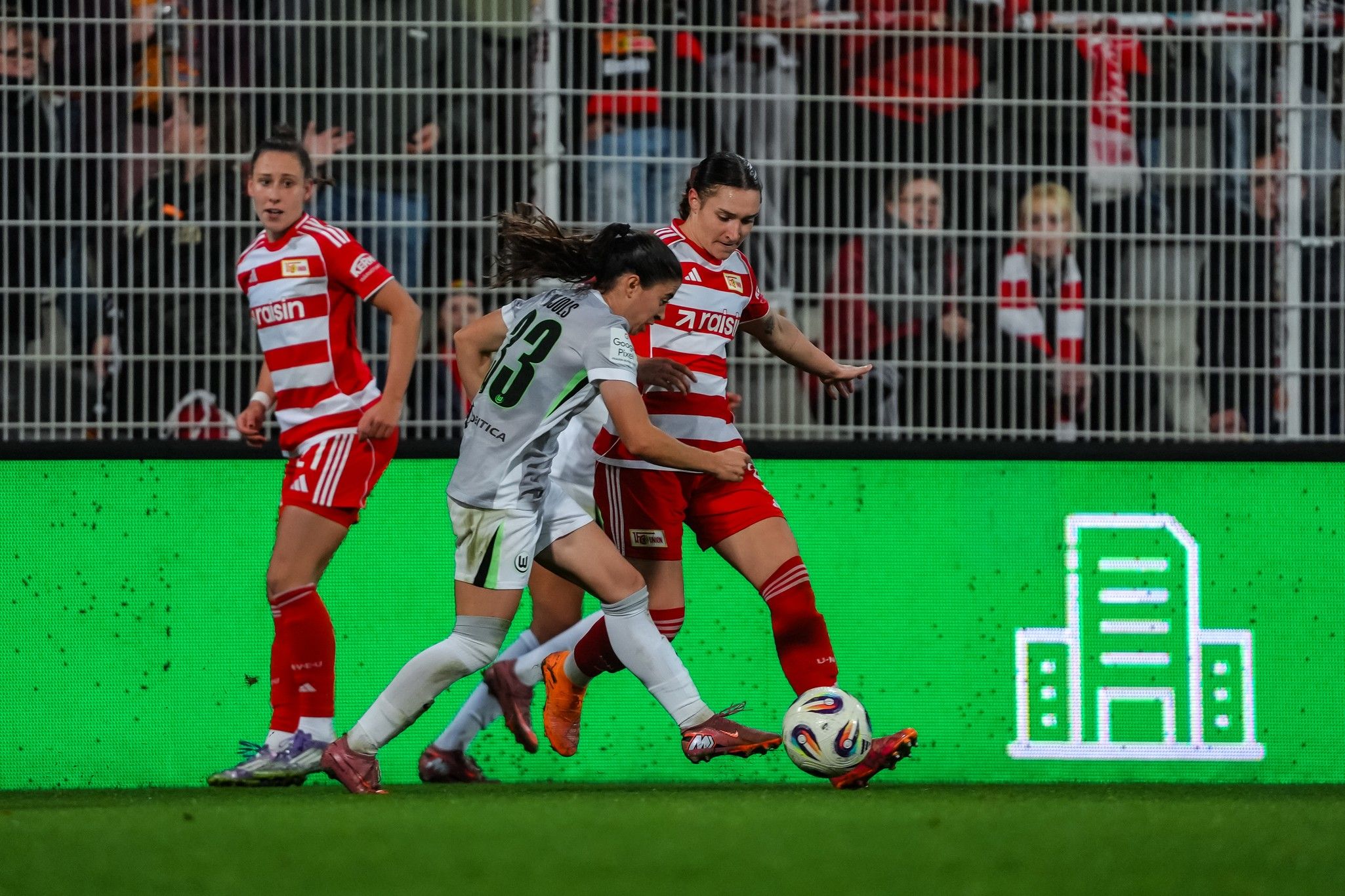 Zwei Fußballspielerinnen kämpfen um den Ball auf dem Spielfeld, während eine weitere Spielerin im Hintergrund agiert.