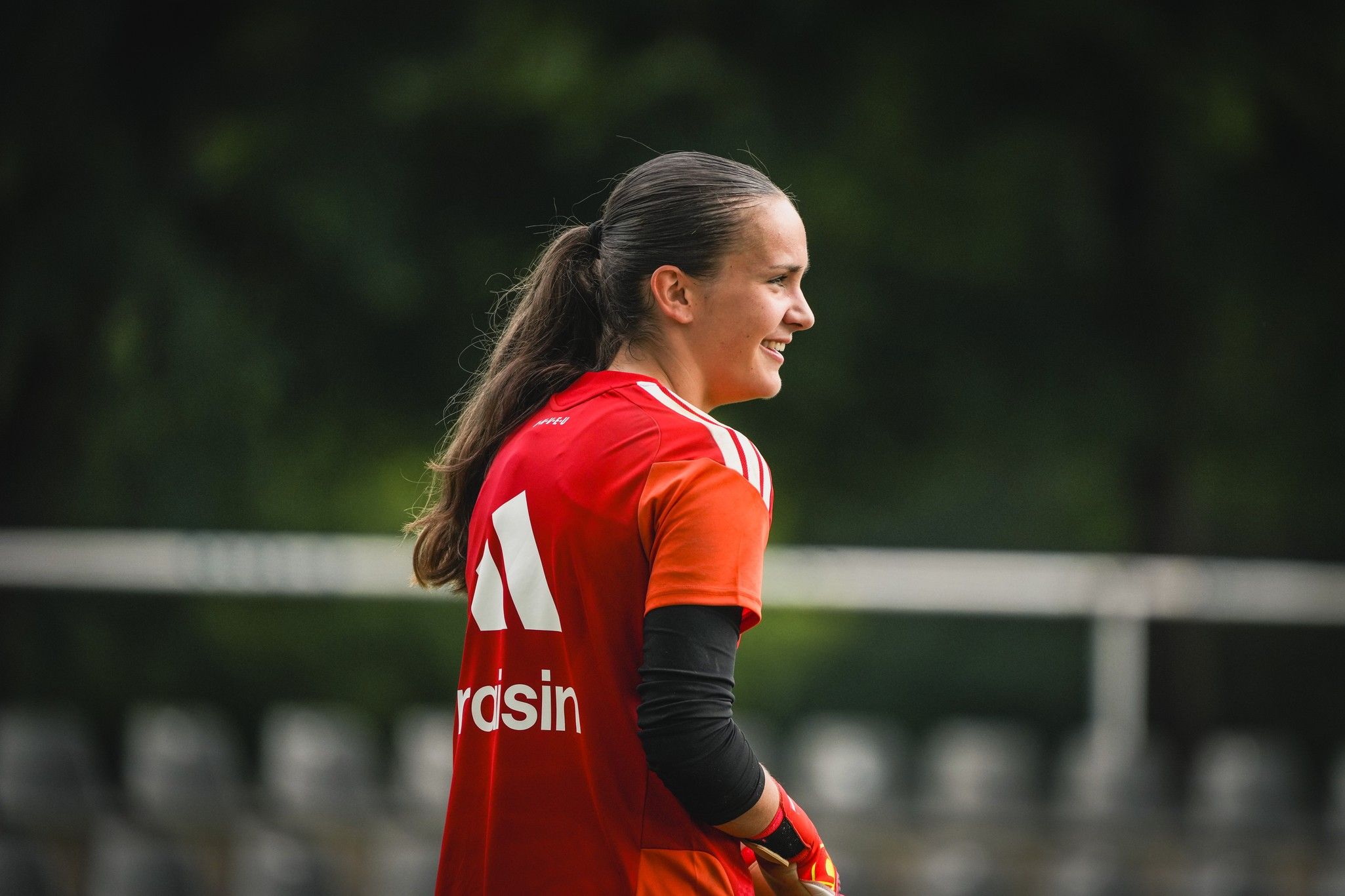Eine junge Torhüterin in einem roten Trikot lächelt, während sie auf einem Fußballplatz steht.