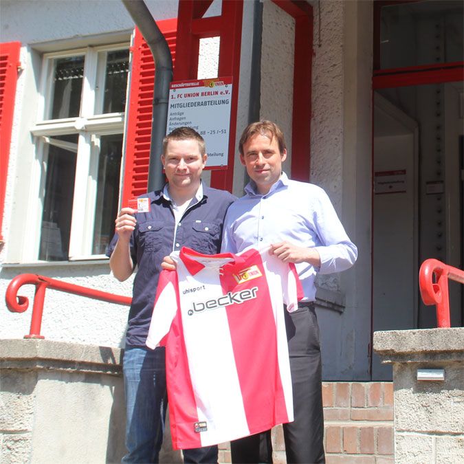 Zwei Männer stehen vor einem Gebäude und halten ein Trikot sowie eine Mitgliedskarte in der Hand.