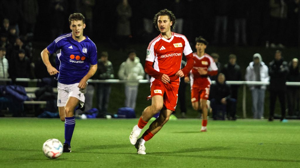 Zwei Fußballspieler spielen auf einem Platz. Viele Zuschauer schauen zu. Es ist Nacht.