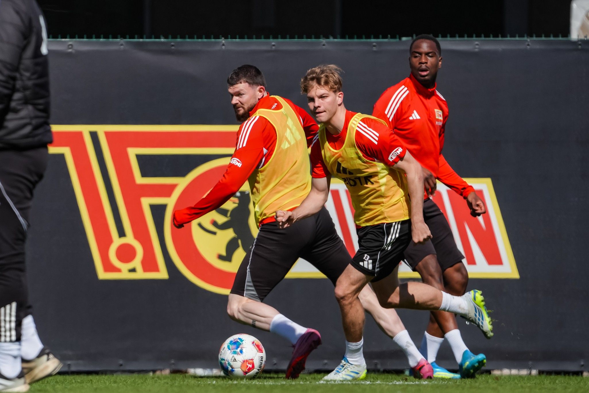 Drei Fußballspieler in Trainingsanzügen absolvieren ein Training auf dem Platz, ein Ball liegt vor ihnen.