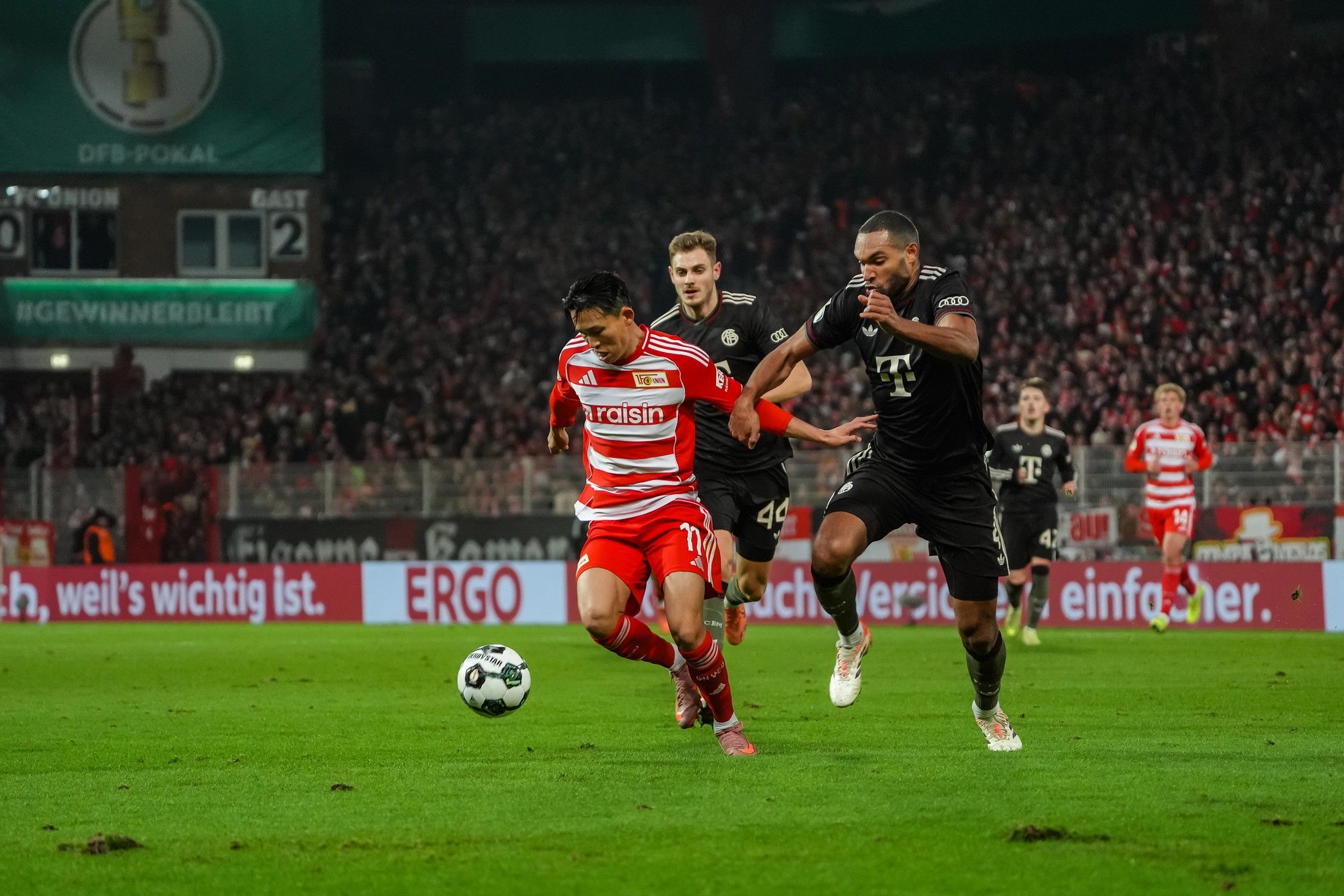 Ein Fußballspieler im roten Trikot mit "Union" Schriftzug rennt auf den Ball zu, während ein Spieler im schwarzen Trikot ihn verfolgt.