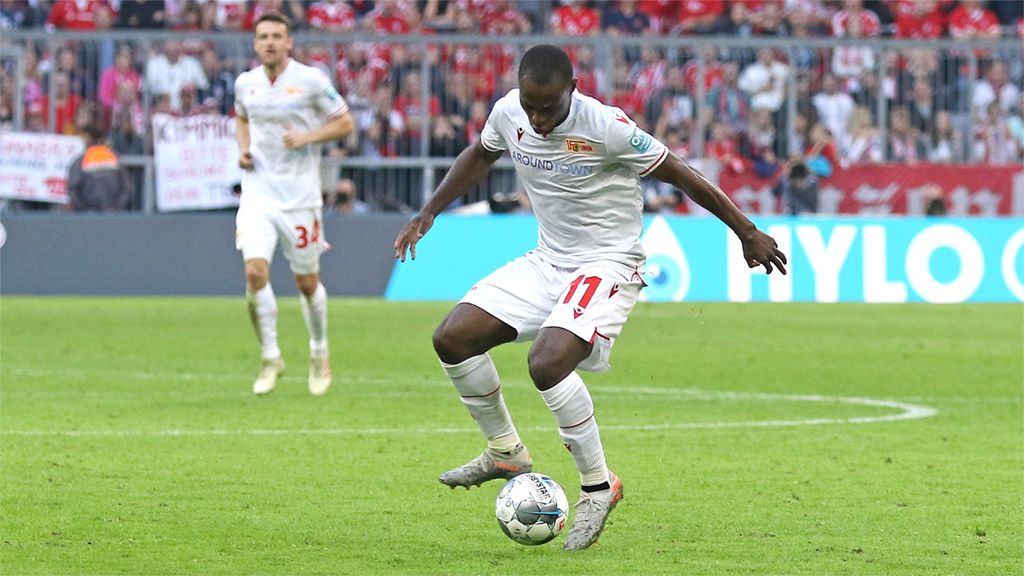 Spieler in weißem Trikot dribbelt den Ball während eines Fußballspiels, im Hintergrund sind Fans und weitere Spieler zu sehen.