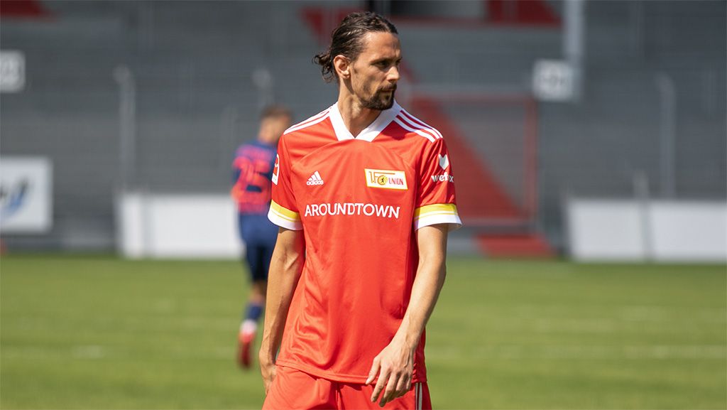 Ein Fußballspieler trägt ein rotes Trikot mit gelben und weißen Akzenten und steht auf einem grünen Rasenfeld.