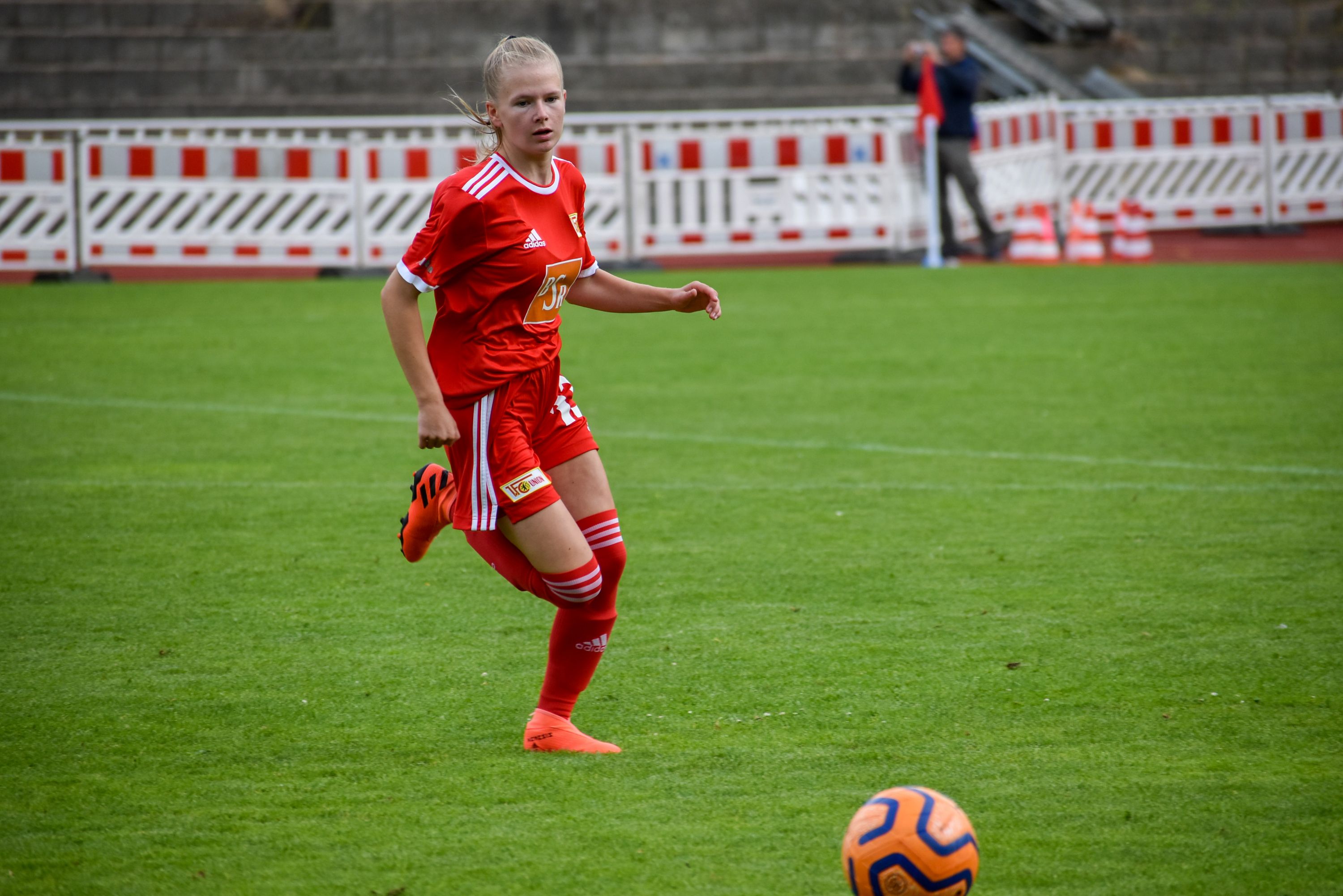 Eine Fußballspielerin in rotem Trikot sprintet über das Spielfeld, der Ball liegt vor ihr auf dem Rasen.
