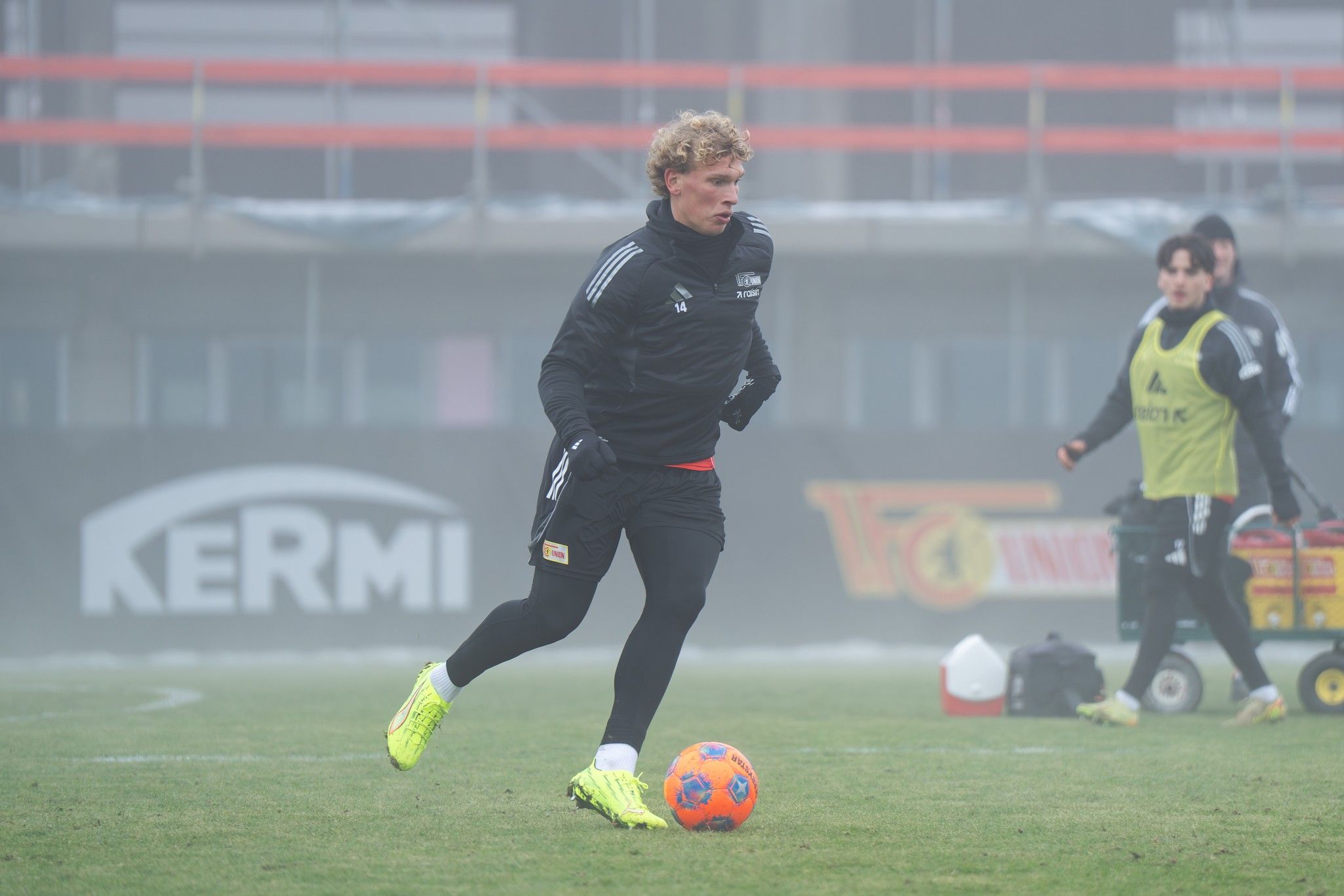 Spieler in dunklem Trainingsanzug mit gelben Fußballschuhen, der auf einem nebligen Fußballplatz dribbelt. Im Hintergrund andere Spieler.
