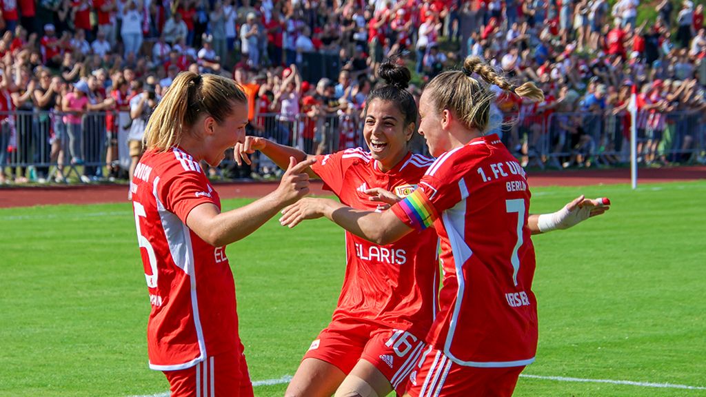 Drei feiernde Fußballspielerinnen in roten Trikots auf dem Platz, umgeben von jubelnden Fans.