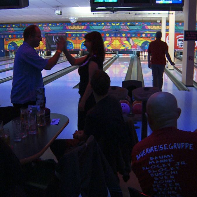 Eine Bowlingbahn mit bunten Wänden, ein Paar gibt sich ein Hoch-Fives, während andere am Tisch sitzen und zuschauen.