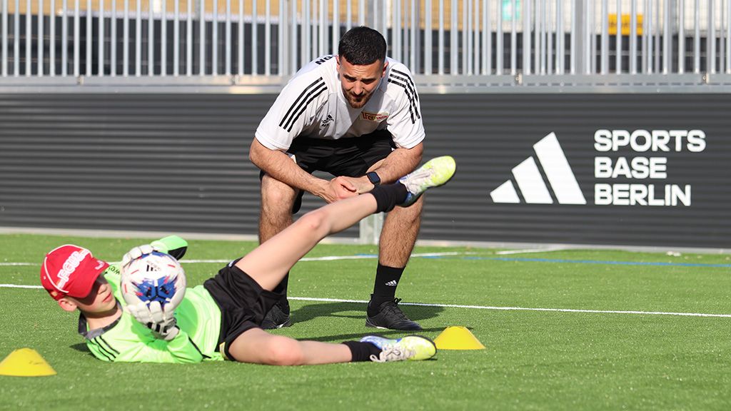 Ein Trainer beobachtet einen jungen Torwart, der beim Training einen Ball fängt. Stangen und Sportplatz im Hintergrund.