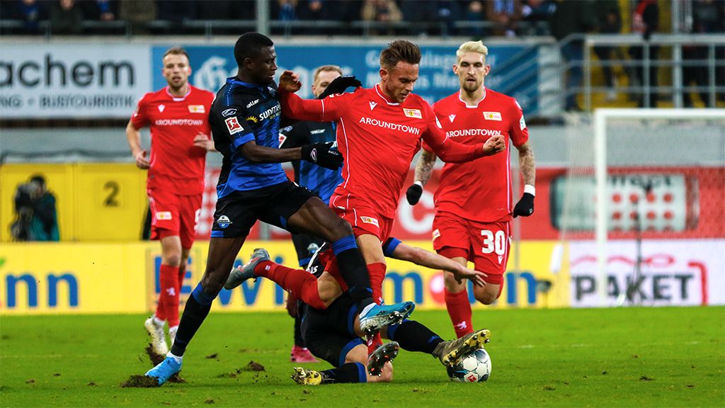Ein Fußballspieler mit rotem Trikot will den Ball. Er kämpft gegen einen Spieler mit blauem Trikot. Sie sind auf dem Feld.