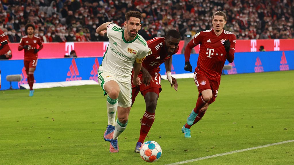 Ein Fußballspieler in weißer Uniform zieht einen anderen Spieler in roter Uniform nach sich, während ein dritter Spieler nahe ist.