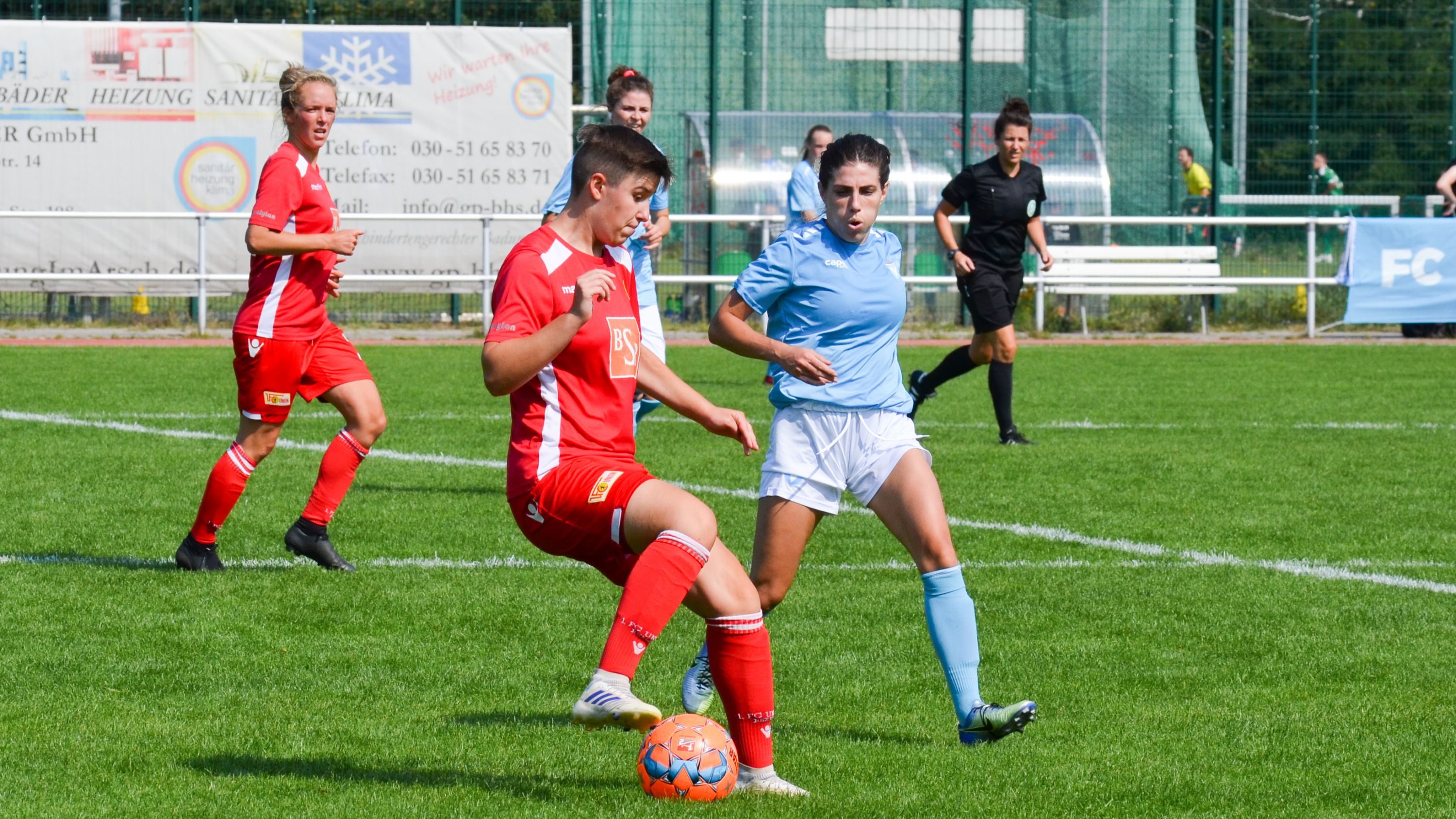Zwei Frauenfußballspielerinnen in roten und blauen Trikots spielen auf einem grünen Feld, während eine Schiedsrichterin im Hintergrund steht.