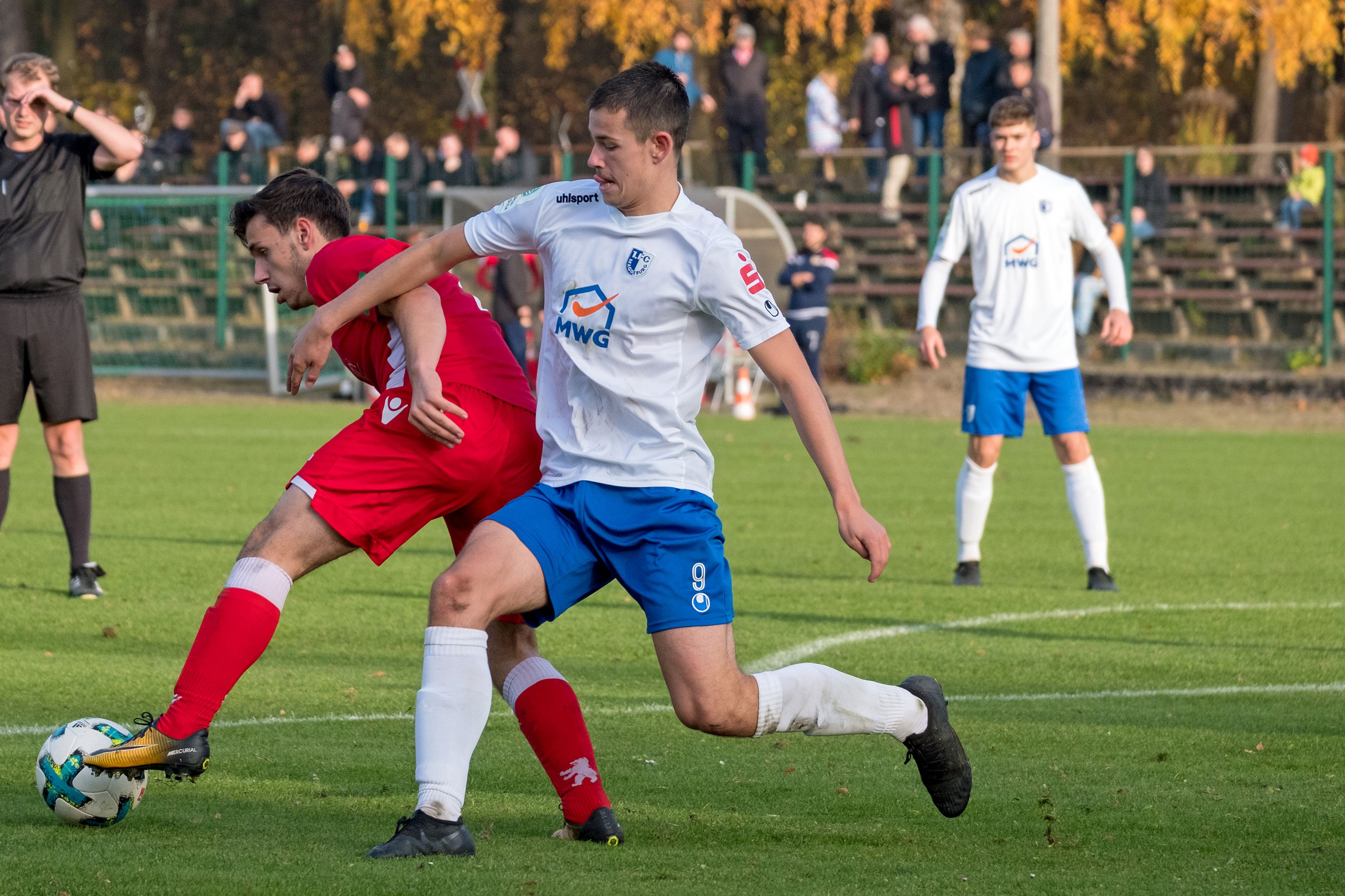 Spielszene eines Fußballspiels, zwei Spieler im Zweikampf, im Hintergrund weitere Spieler und Zuschauer. Herbstliche Szenerie.