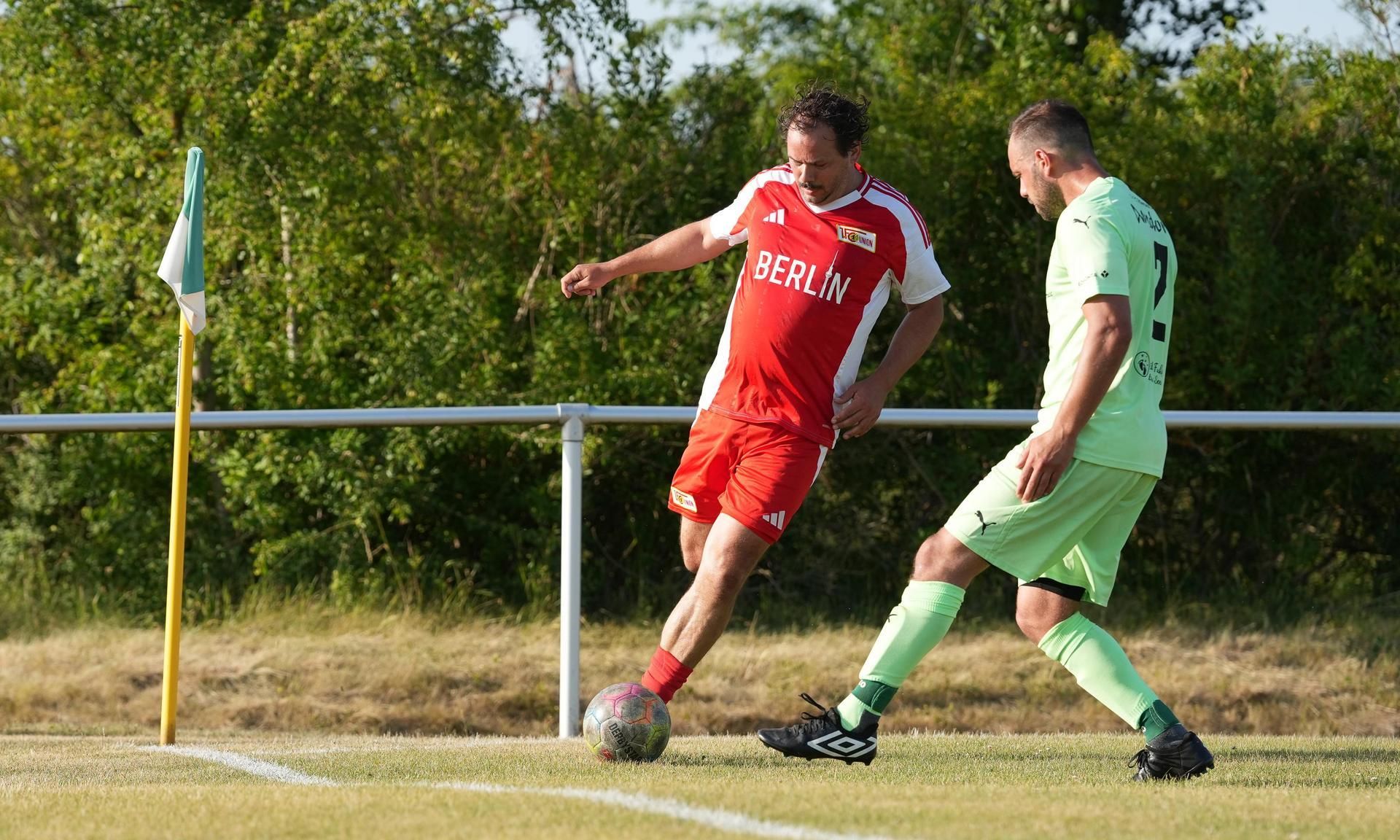 Ein Fußballspieler in rotem Trikot mit der Aufschrift "BERLIN" tritt den Ball, während ein gegnerischer Spieler in grünem Trikot daneben steht.